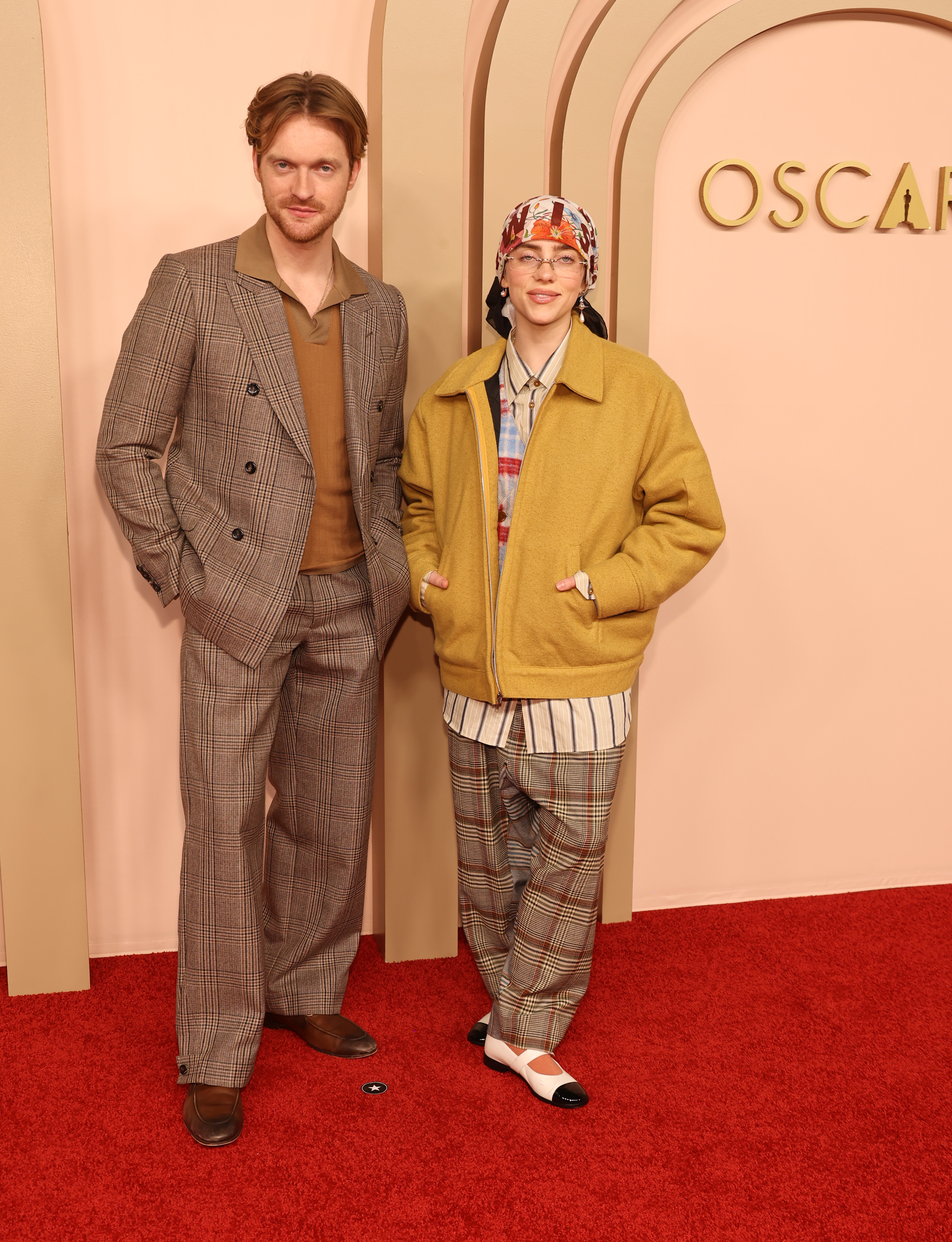 Finneas and Billie standing in front of an &quot;OSCARS&quot; backdrop. Finneas is wearing a checked suit