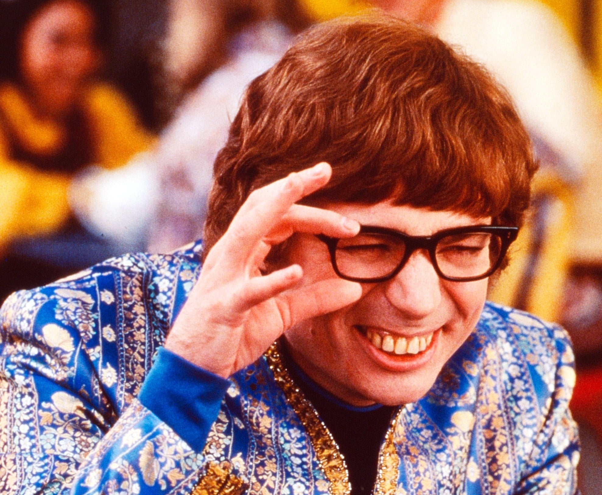 Person in a patterned blue outfit with glasses making a hand gesture near eyes