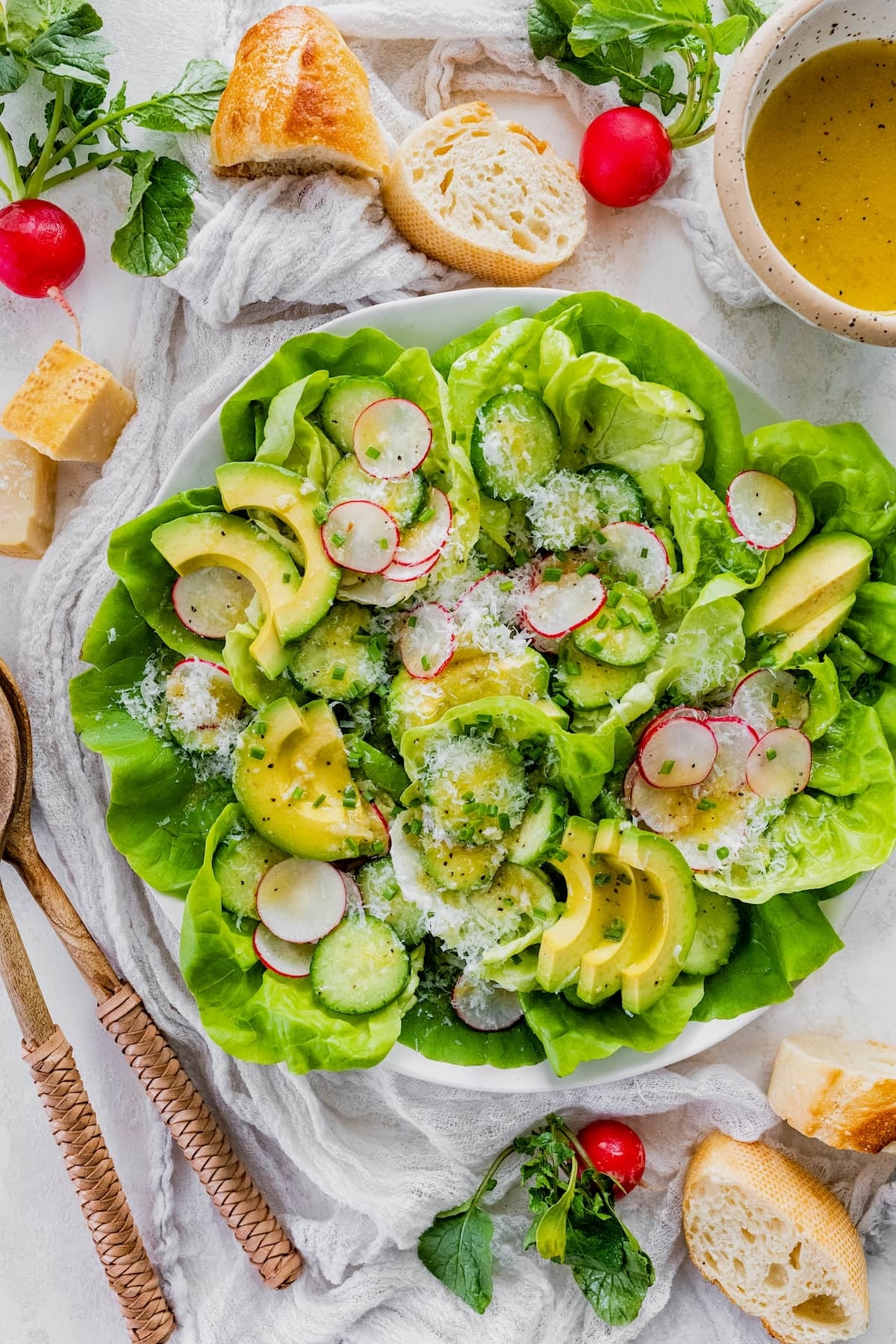 Fresh salad with avocado slices, radishes, cucumber, lettuce, and grated cheese, served with bread on the side