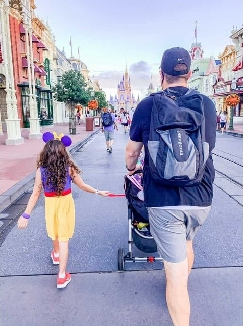 A man and child walk down a theme park&#x27;s main street; the child wears mouse ears and the man has a backpack