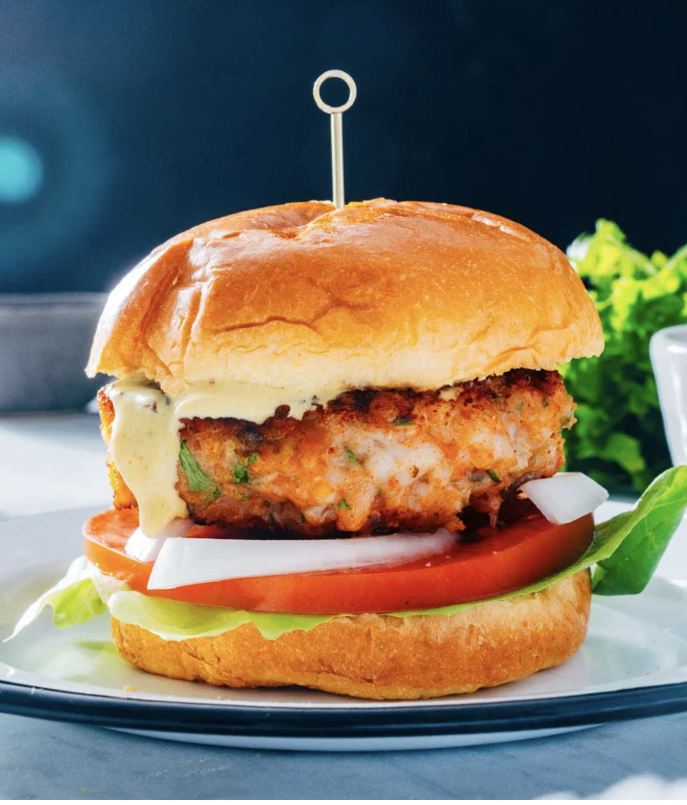 Burger with a crispy chicken patty, lettuce, tomato, and sauce on a brioche bun, skewered with a toothpick