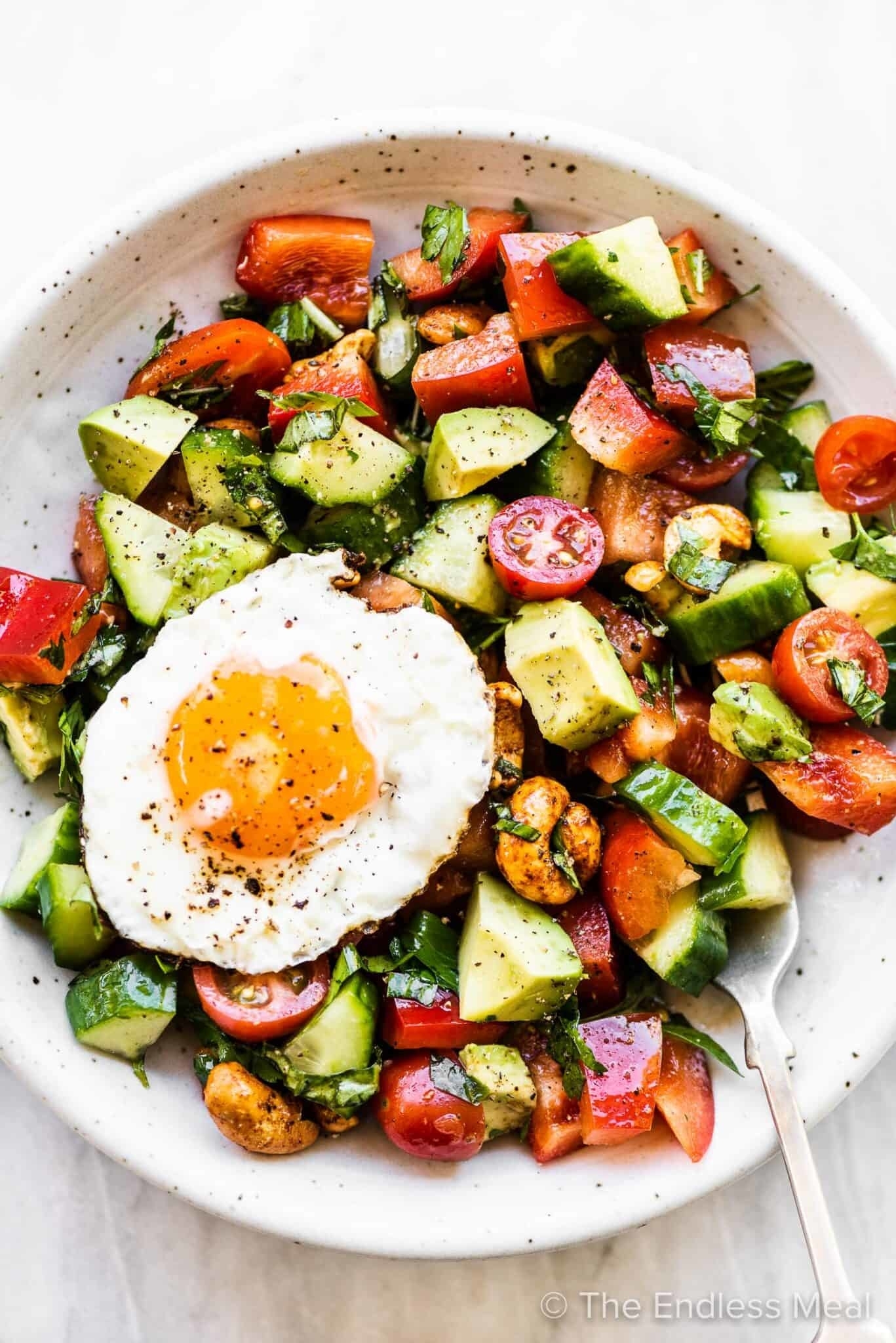 A bowl of salad with tomatoes, cucumbers, avocado, and a fried egg on top, with a fork on the side