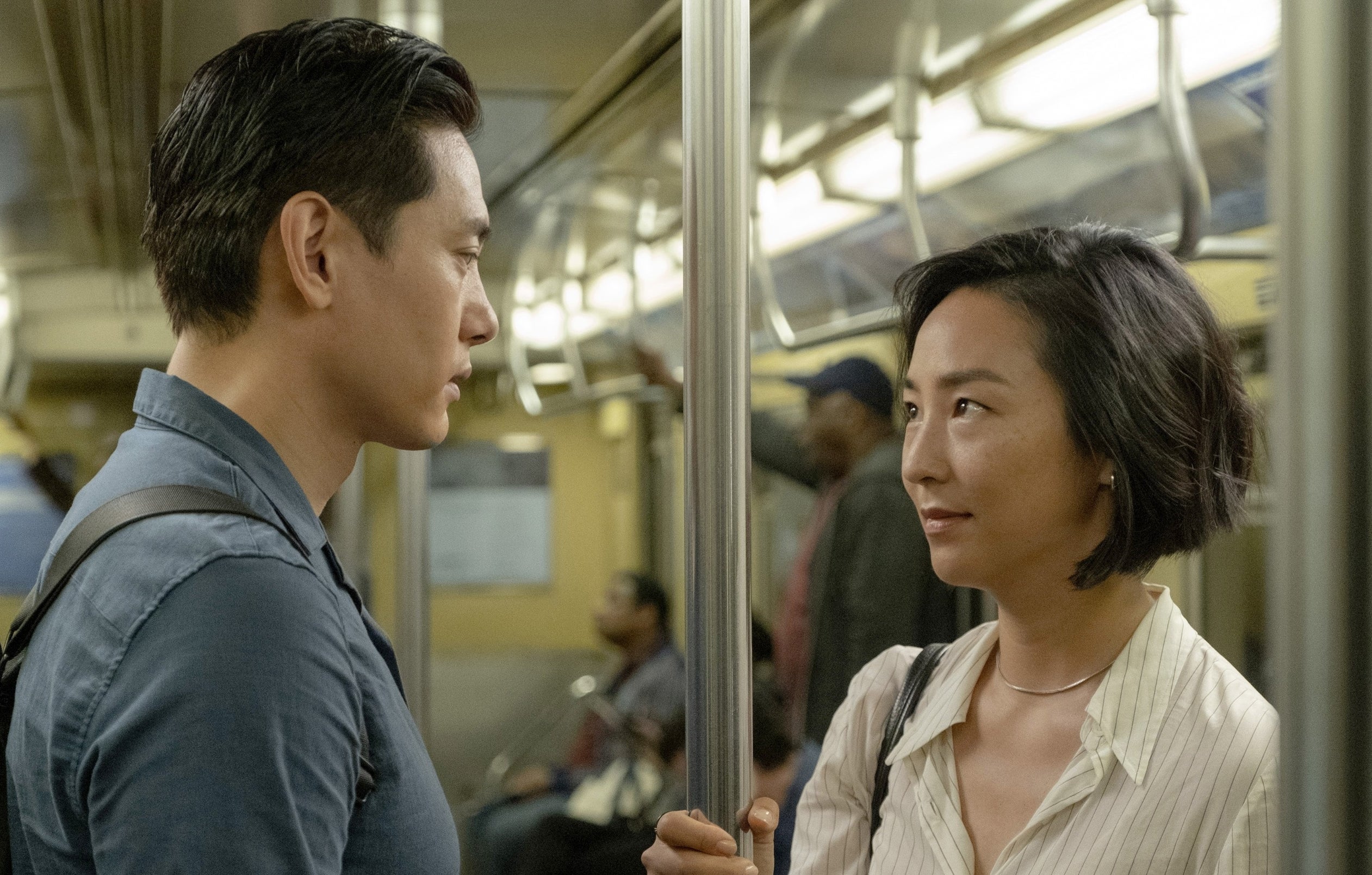 Greta Lee) and Teo Yoo in a subway scene, standing and facing each other, with a pole between them