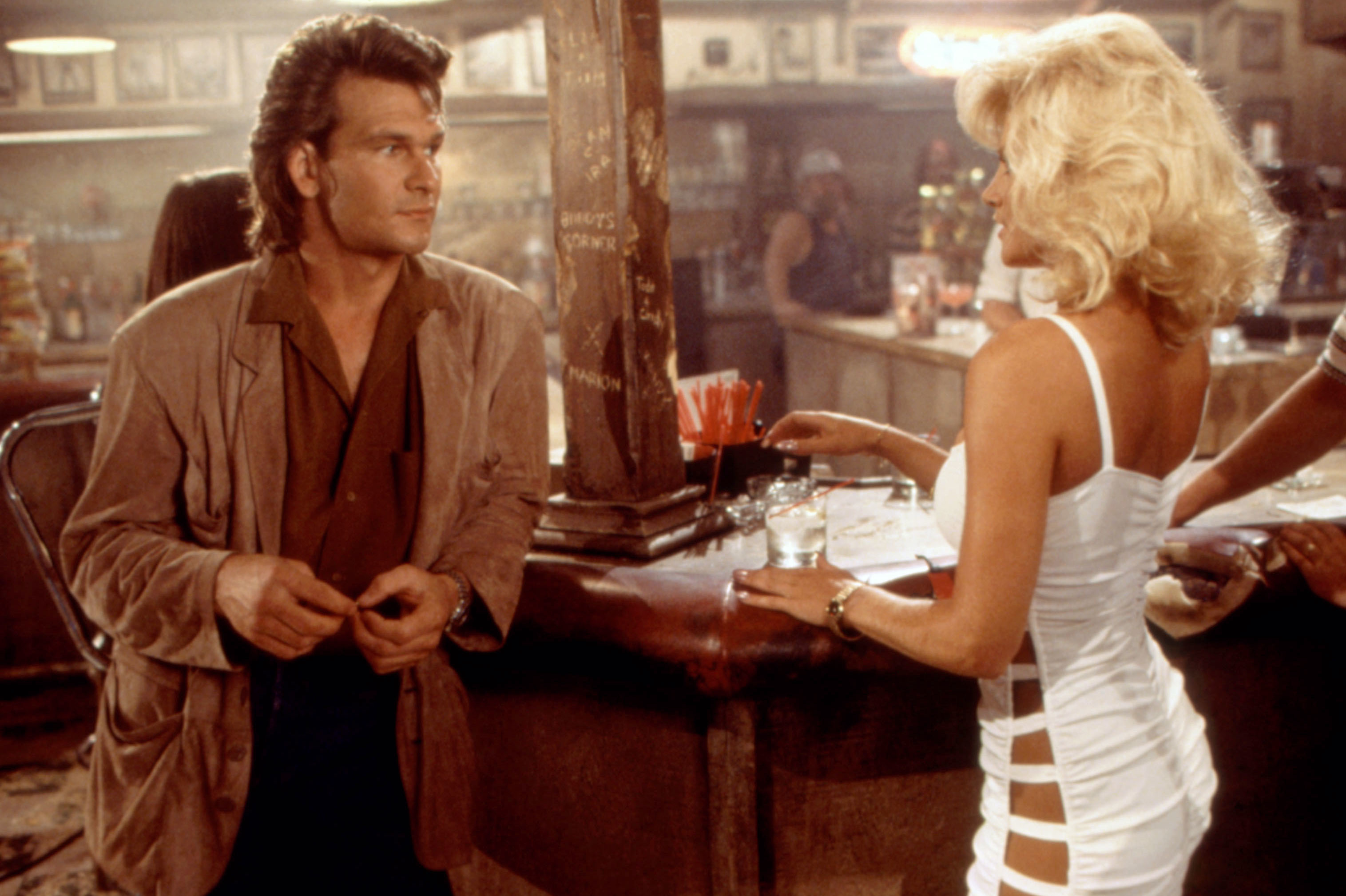 Patrick Swayze, in a suede jacket, speaks to a blonde woman in a skimpy white dress, at a bar