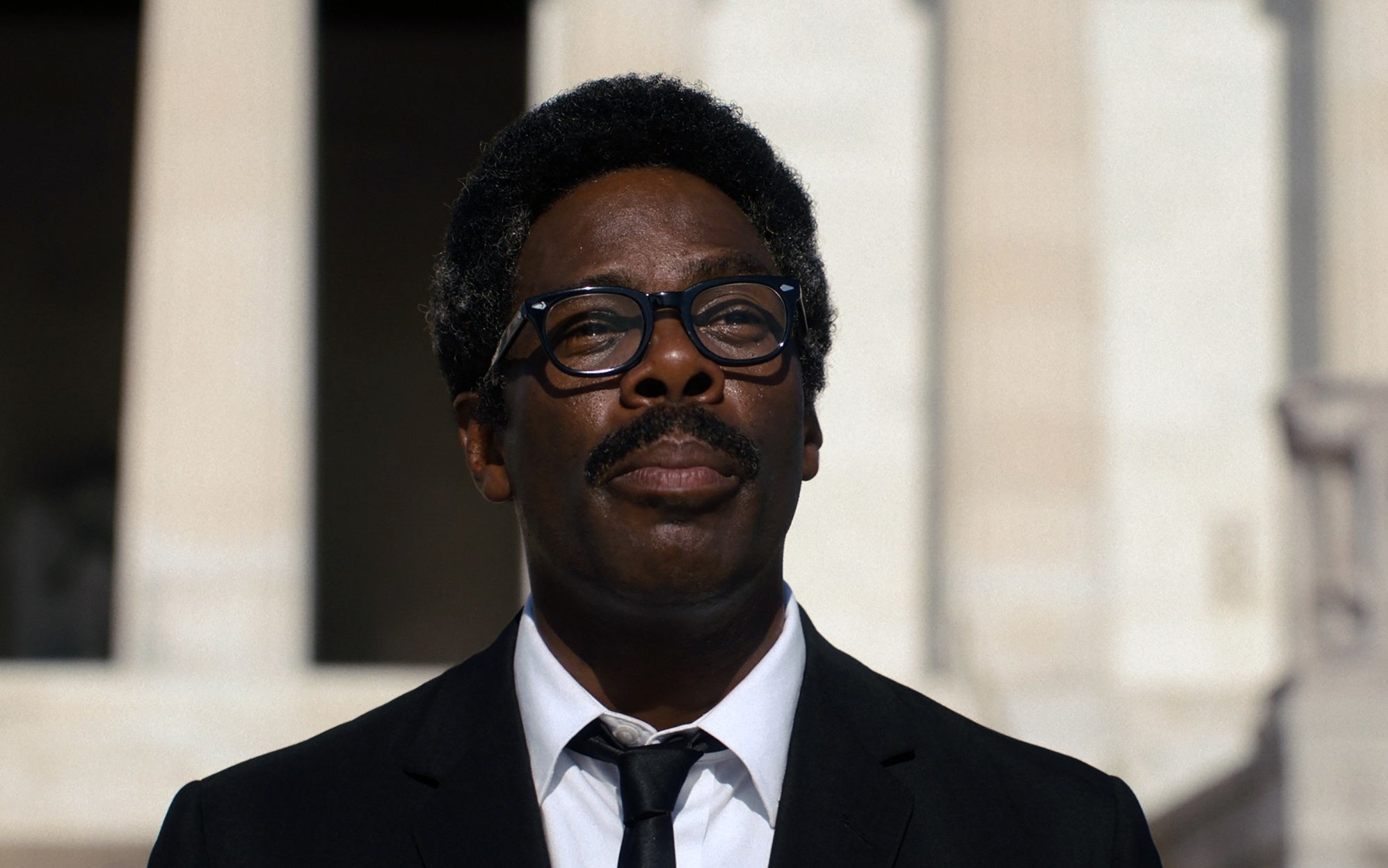 Colman Domingo in a suit and tie with glasses standing in front of a building, expression neutral