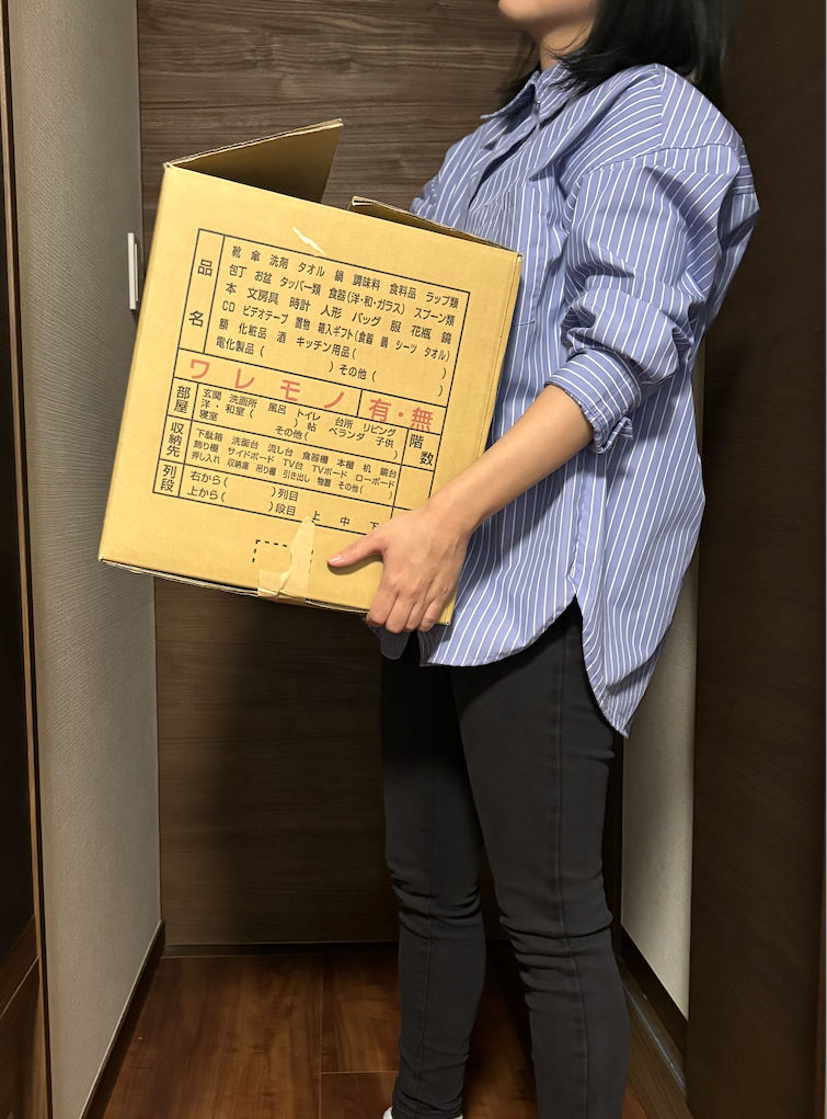 ストライプシャツを着た人が段ボール箱を持って立っています。箱には多くの日本語テキストが含まれています。