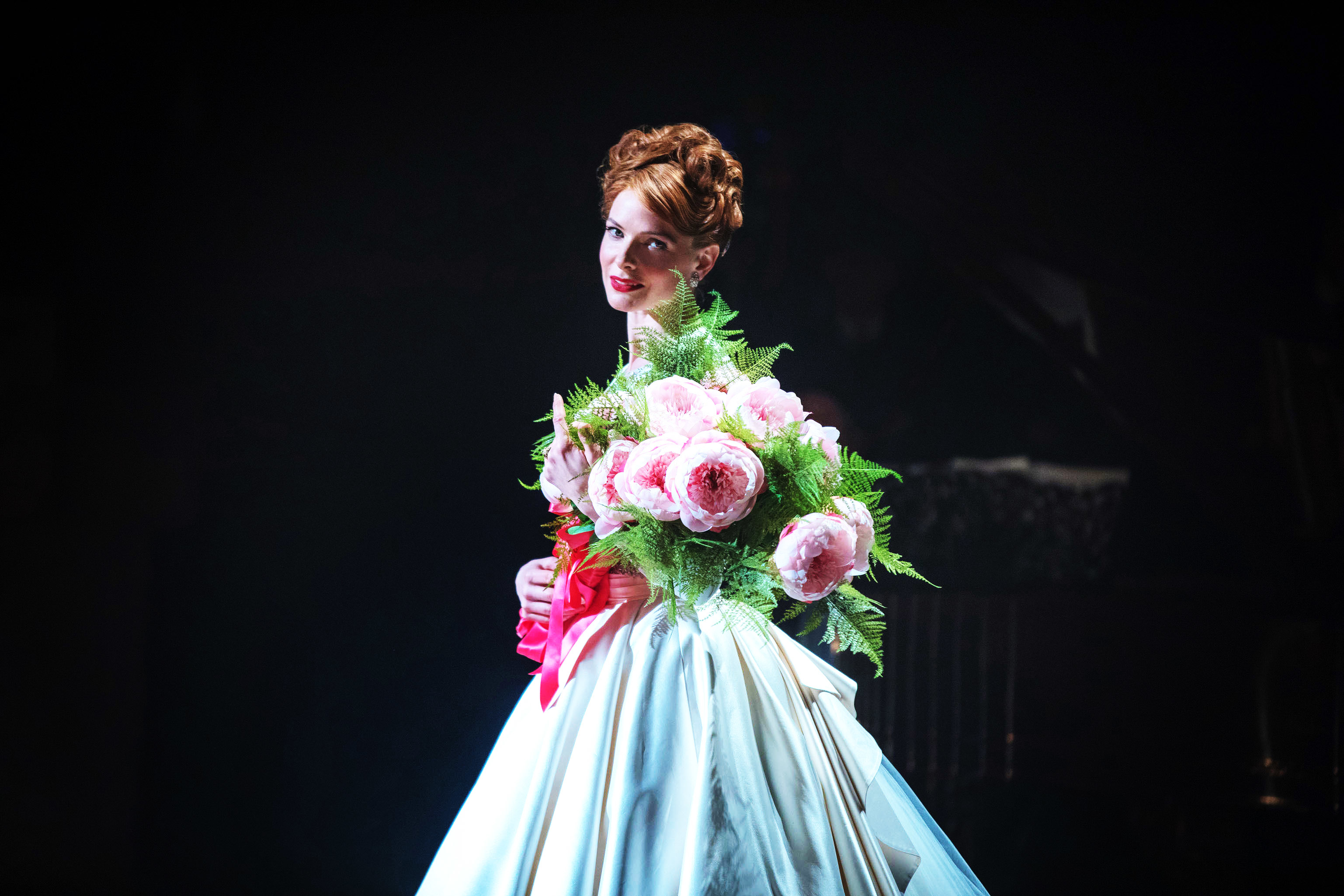 Rebecca Ferguson in historical costume holds bouquet onstage, evoking a classical period drama scene