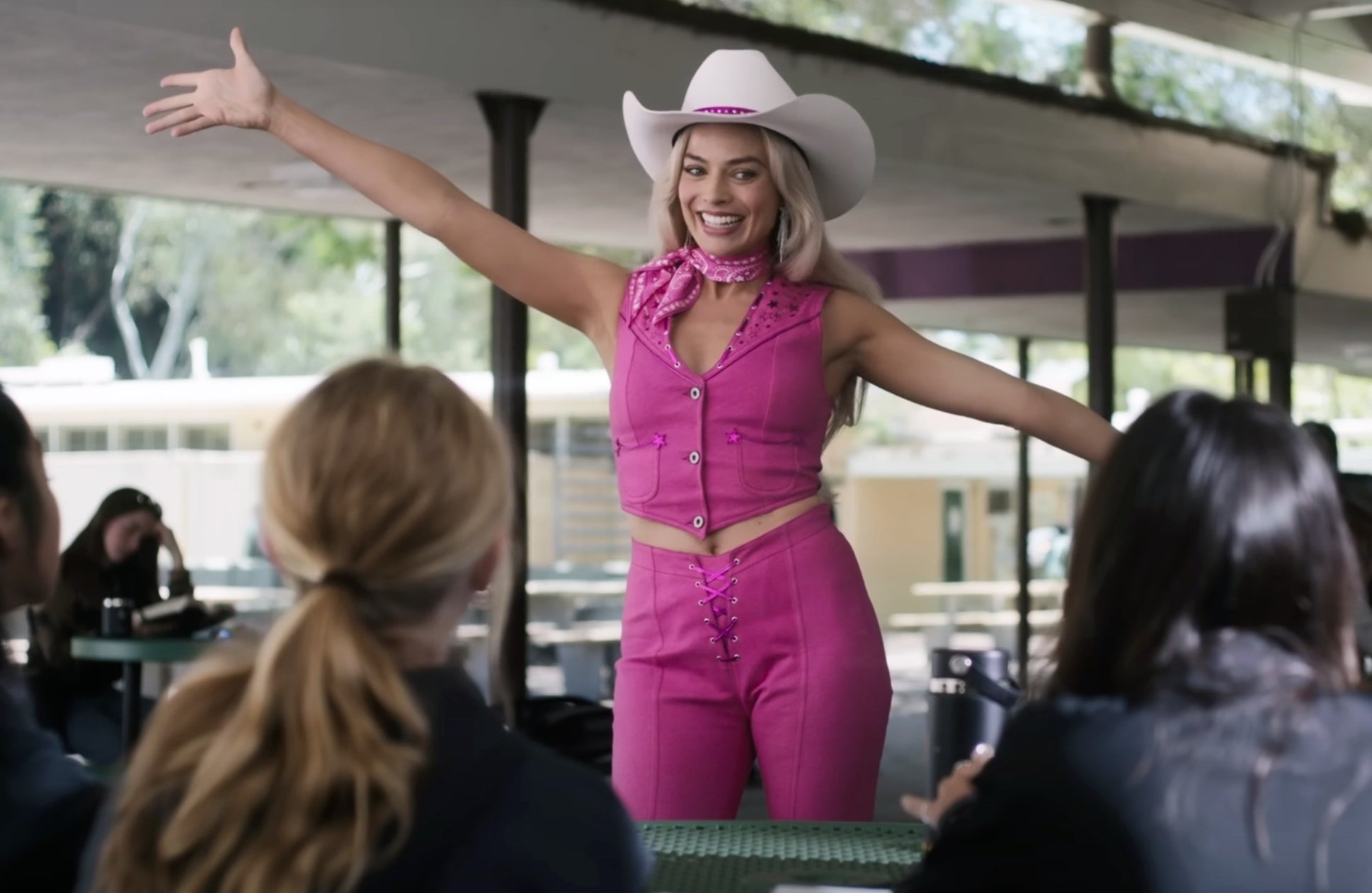 Margot Robbie in a cowboy hat and pink ruffled outfit gesturing with arms wide, standing indoors with others around