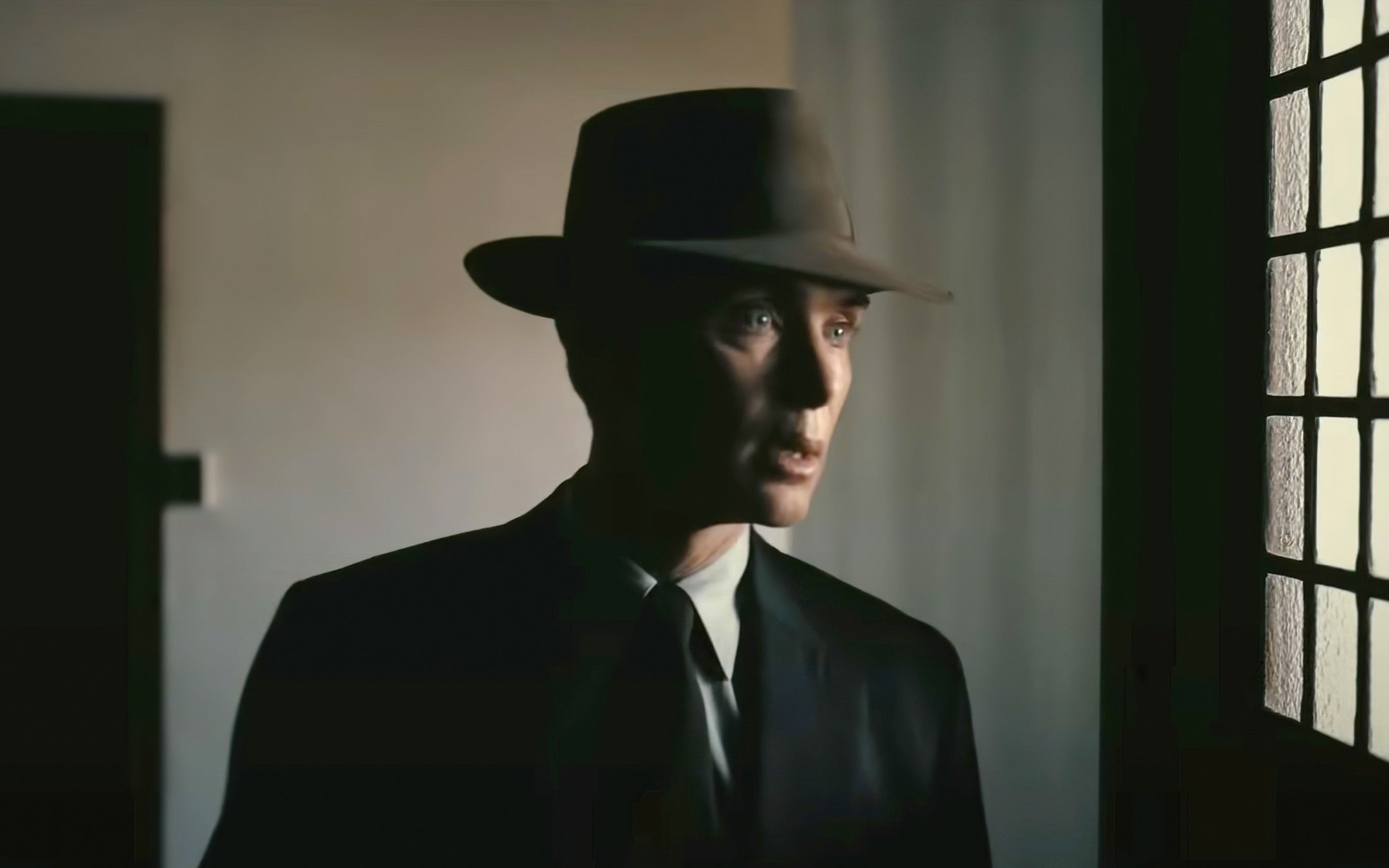 Cillian Murphy in a vintage suit and fedora hat stands in a dimly lit room