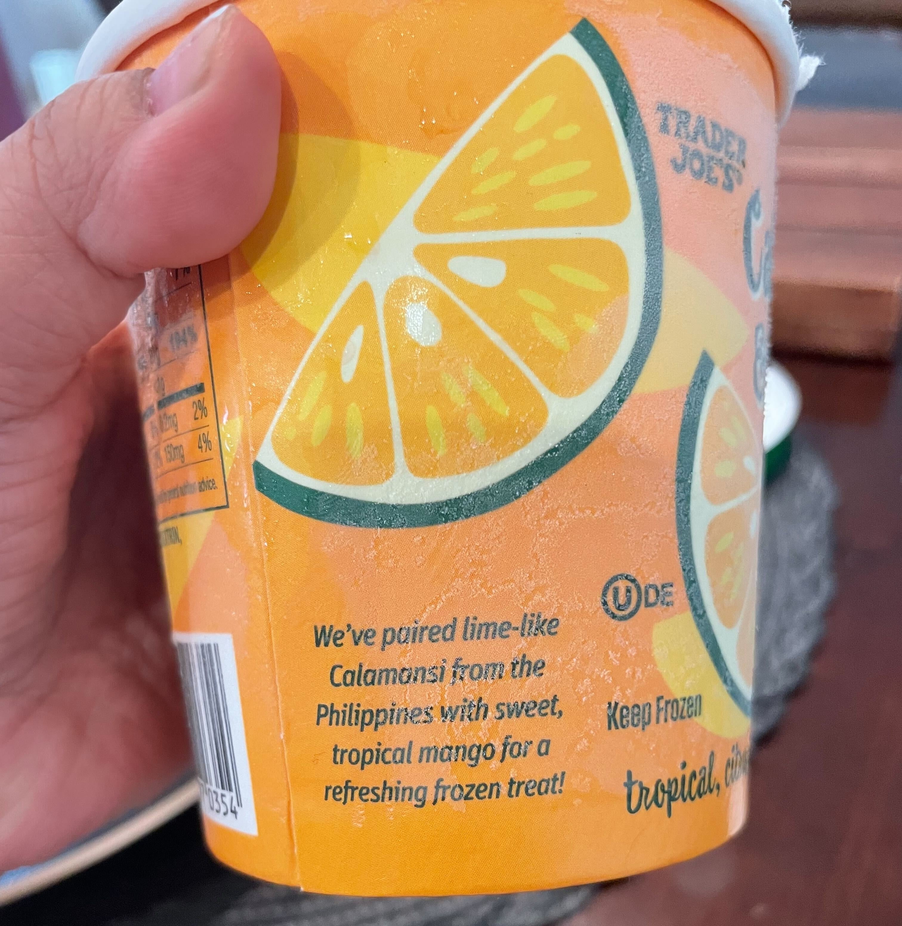 Hand holding a Trader Joe&#x27;s frozen treat with lime and calamansi fruit graphics. Text: &quot;We&#x27;ve paired lime-like Calamansi from the Philippines with sweet, tropical mango for a refreshing frozen treat!&quot;
