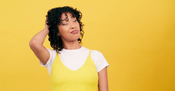 Person posing with hand in hair wearing a yellow top and white tee, appears contemplative, against a yellow backdrop