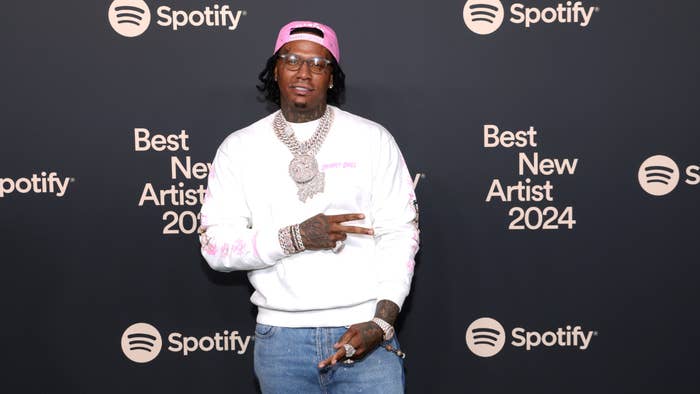 Rapper at Spotify event, wearing sweater and chains, smiling in front of promotional backdrop