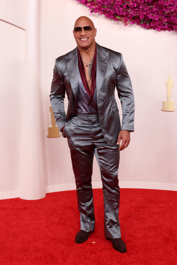 Dwayne Johnson in a shiny gray suit with a deep red shirt, posing on the red carpet