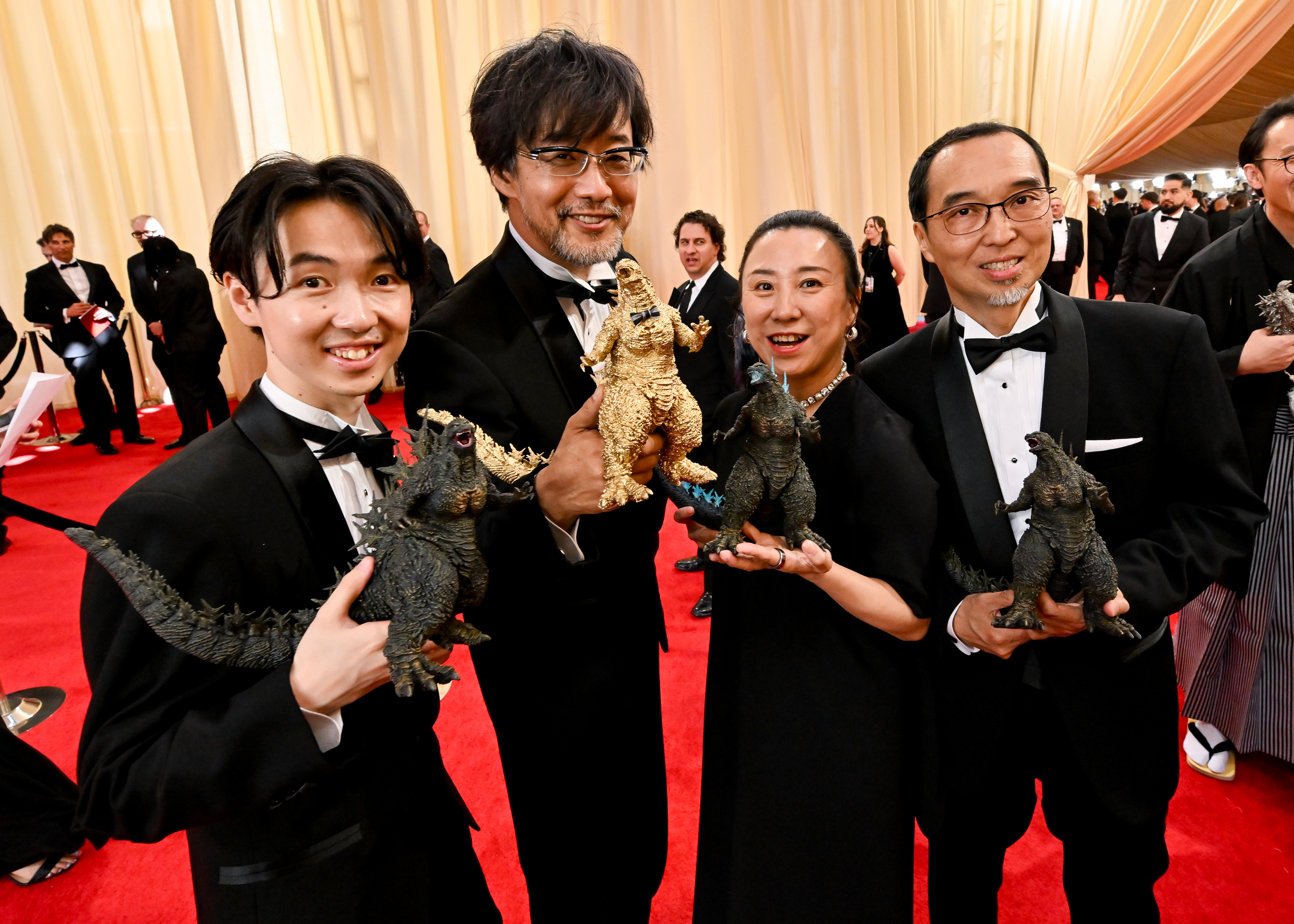 Four individuals posing with Godzilla figures; two men in tuxedos, one woman in dress, celebratory event