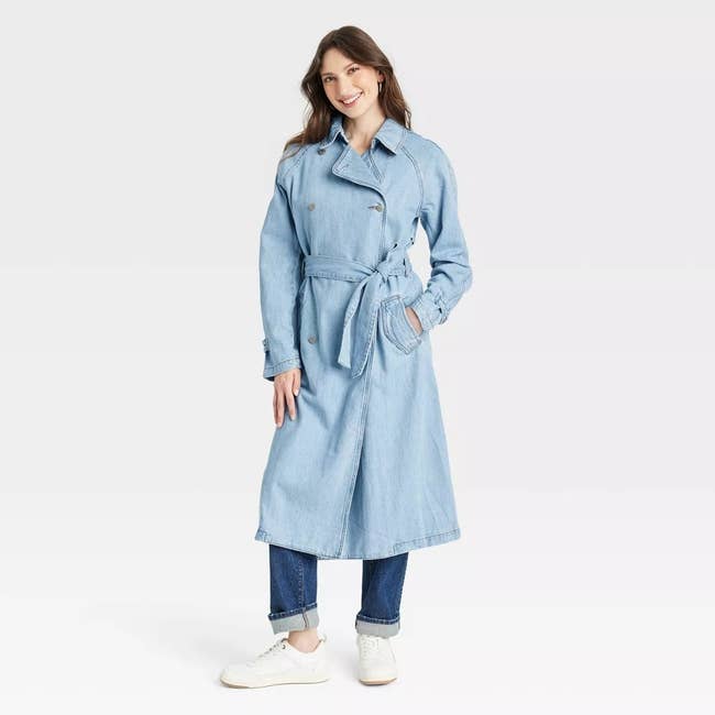 A person models a long denim trench coat with a belt