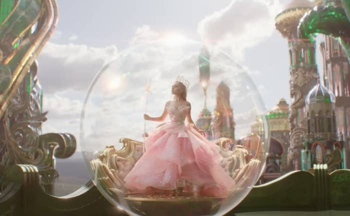 Ariana Grande in an elaborate pink dress sits within a transparent bubble in front of a fantastical palace background
