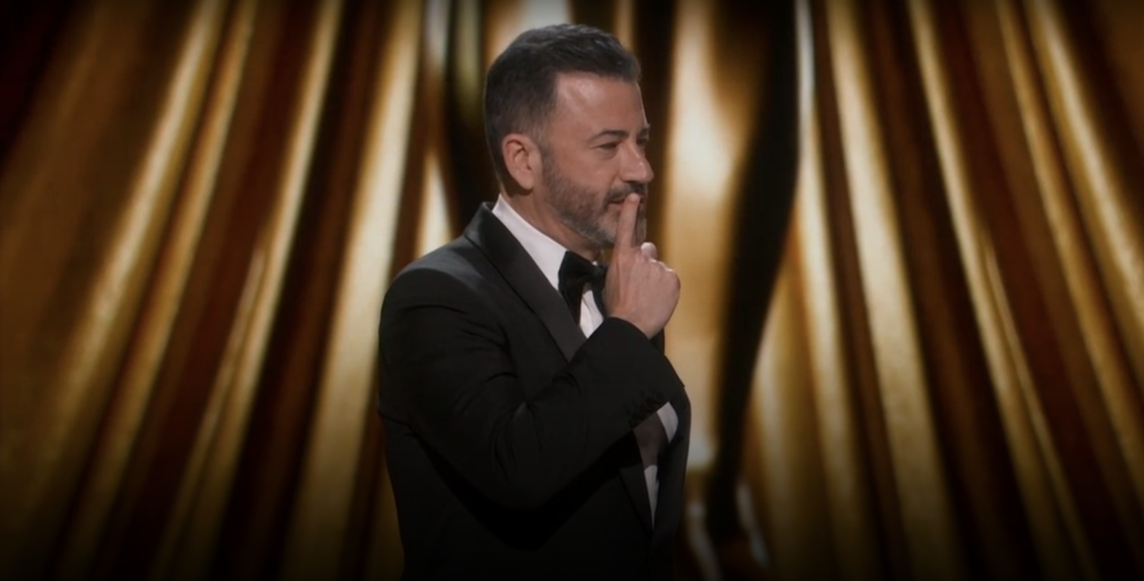 Jimmy Kimmel in a tuxedo hosting on stage with a thoughtful expression, finger on chin