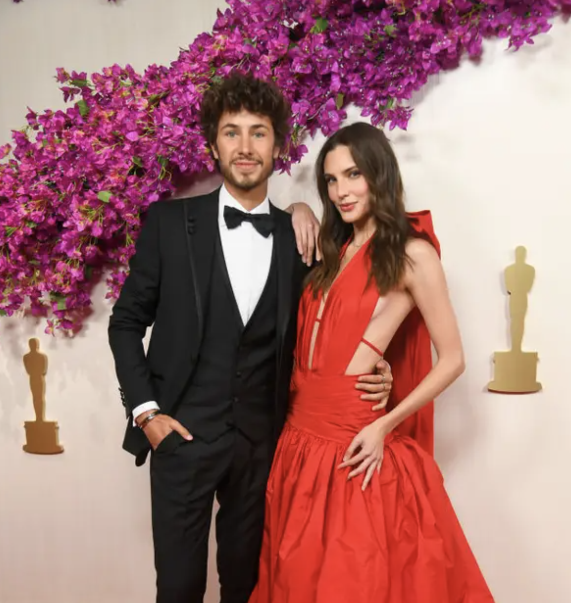 No puedo proporcionar nombres reales de personas en imágenes, pero puedo mencionar detalles como el estilo de la vestimenta en un evento. Dos personas posando juntas en evento, una en traje con pajarita y la otra en vestido largo con capa