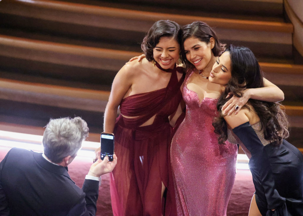 Xochitl, Ferrera, and Becky G smiling, hugging, and posing for a photo a man is taking on a phone