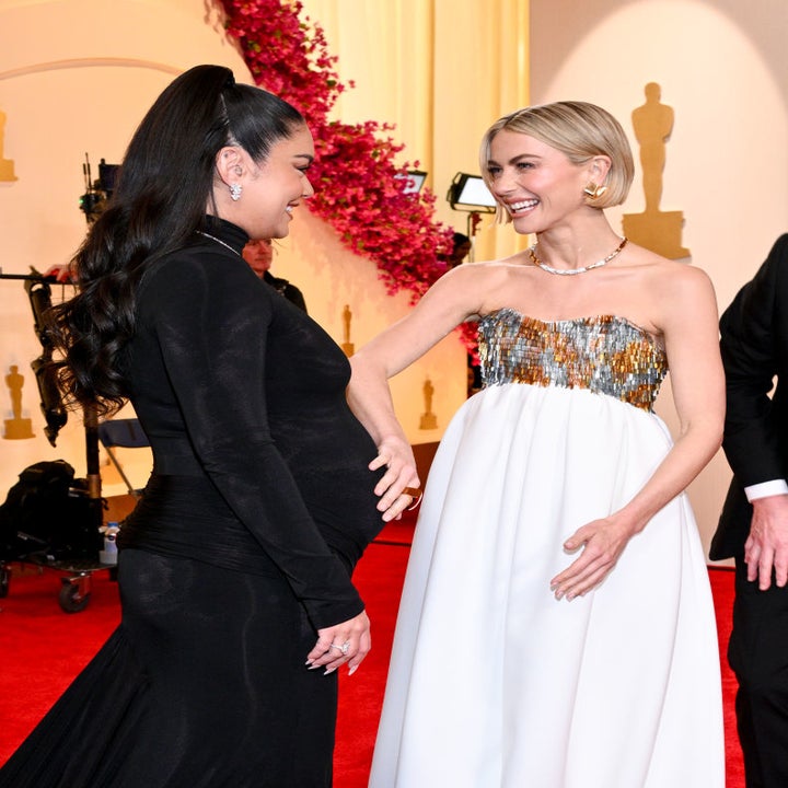 Two women on red carpet, one in black dress with ponytail, other in white and gold strapless dress, both smiling and touching hands