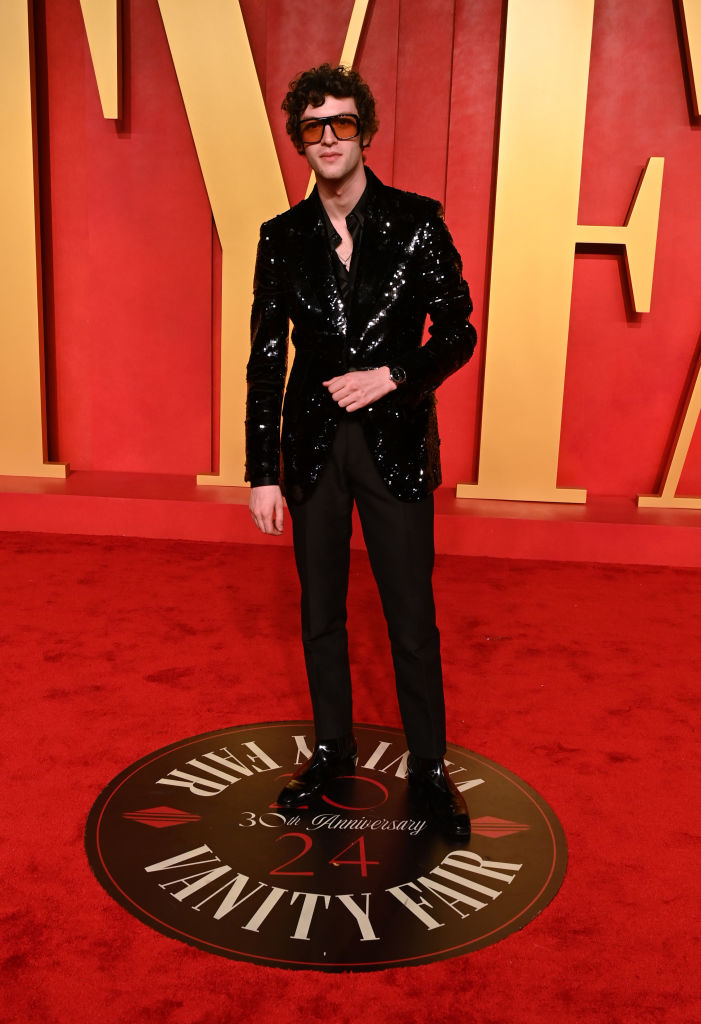 Dominic on red carpet wearing a sequined black suit and curly hair, standing on Vanity Fair logo