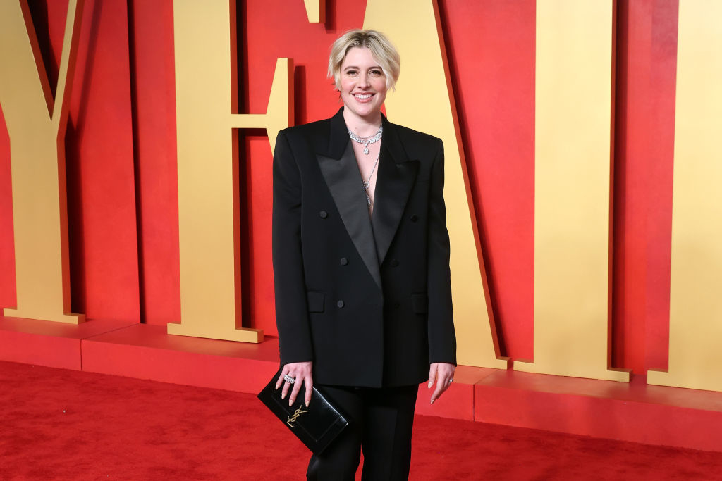 Greta in a black pantsuit and holding a clutch stands smiling on the red carpet
