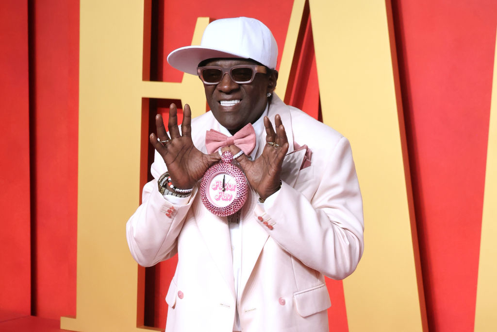 Flavor posing with hands near face, wearing a wide-brimmed cap, suit with bow tie, and large clock necklace