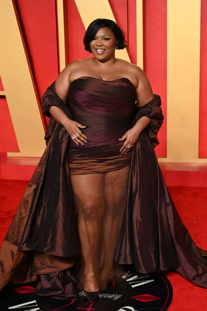 Lizzo in an elegant strapless minidress with sheer long bottom and matching cape, posing on the red carpet