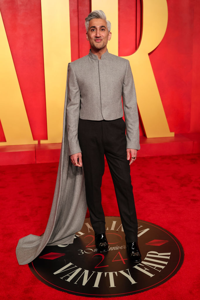 Tan in a gray high-neck top with a long flowing train, black trousers, and polished shoes stands on the Vanity Fair red carpet