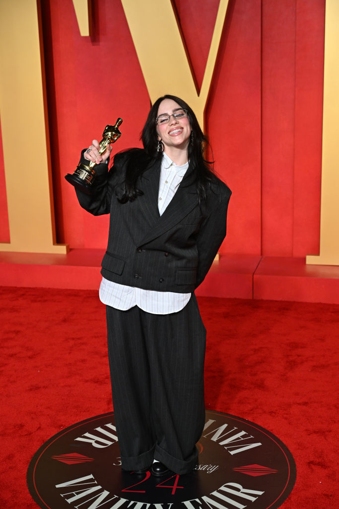 Billie in an oversize pin-striped pantsuit holding an award on a red carpet