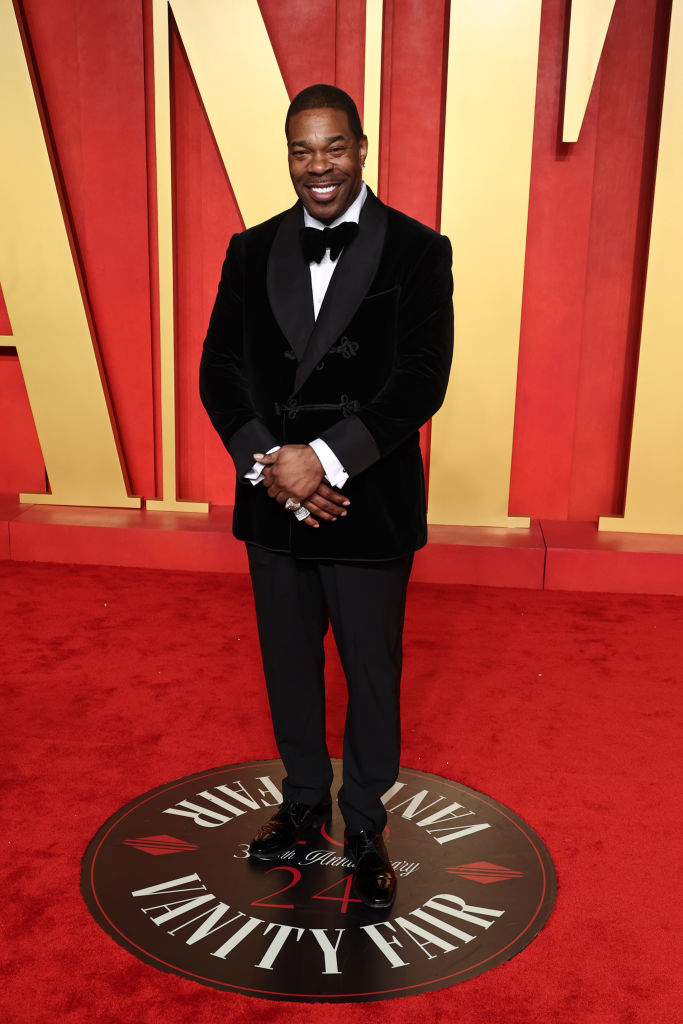 Busta in a black tuxedo with bow tie, smiling, standing on a Vanity Fair emblem at an event