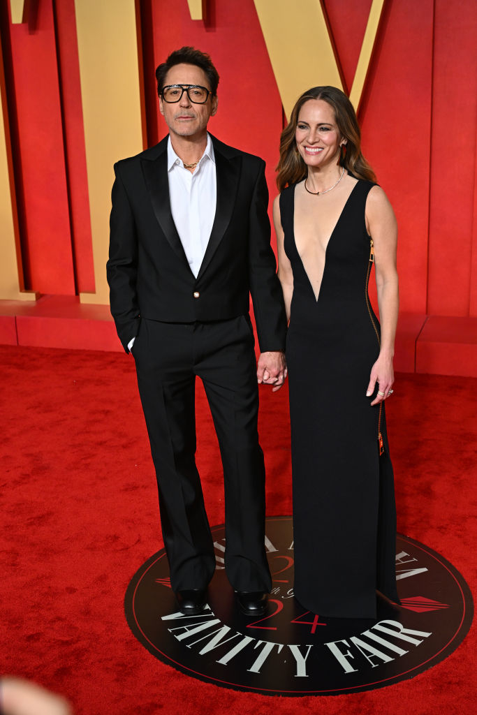 Robert and Susan standing together and holding hands; he&#x27;s in a classic suit, she&#x27;s in a black evening gown with a plunge neckline