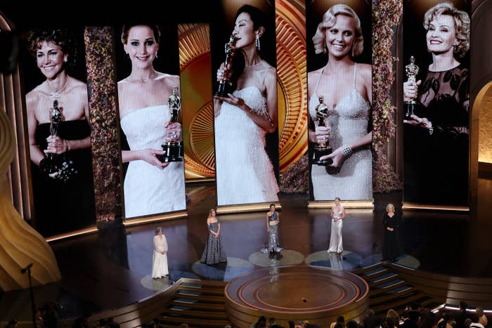 Five women onstage at an awards ceremony, with their images onscreen behind them showing their past wins
