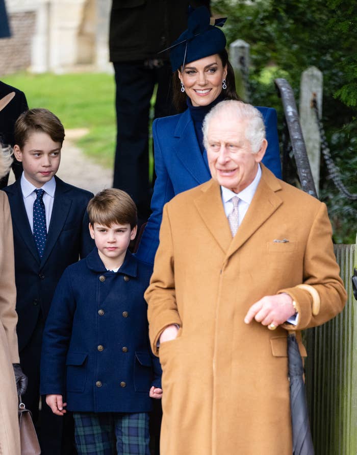 Kate in a blue coat and hat smiles behind her two sons in dark coats and King Charles III in front in a tan coat