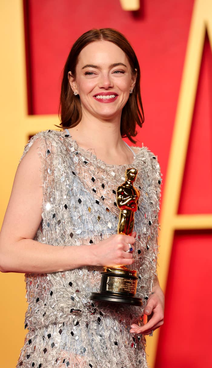 Emma in a beaded dress holding an Oscar statuette and smiling