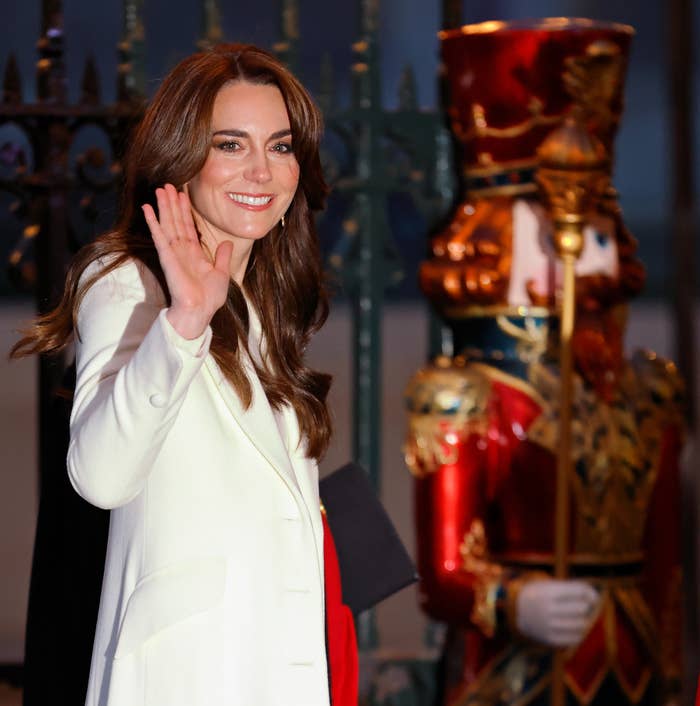 Kate waves, standing by ornate soldier figure; she wears a white coat and smiles