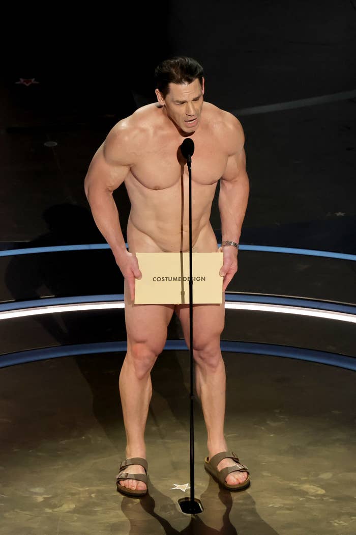 John Cena onstage at an award show, wearing only a small modesty garb and sandals, holding an award category envelope