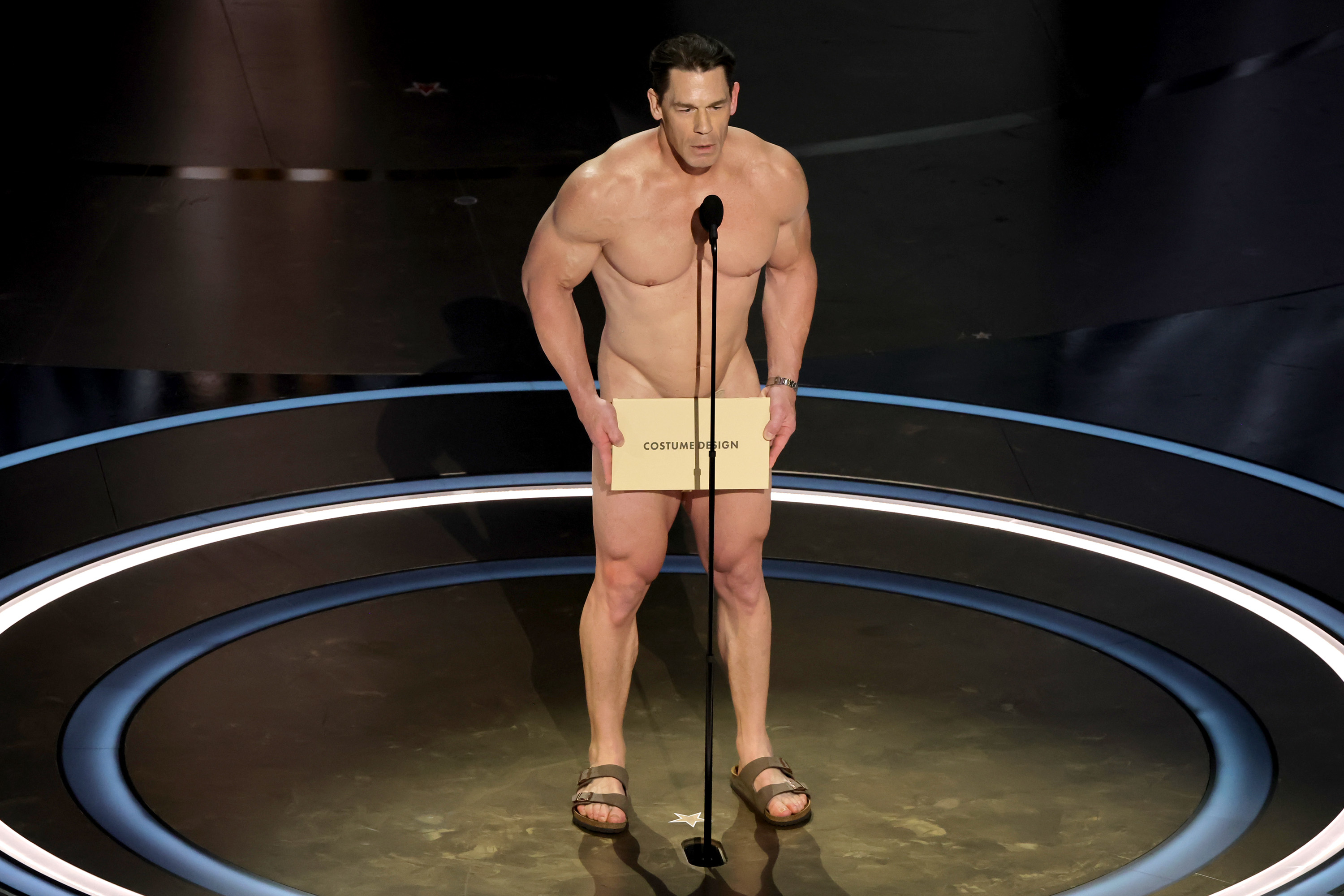 John Cena stands shirtless at a microphone on a stage with a modesty cover