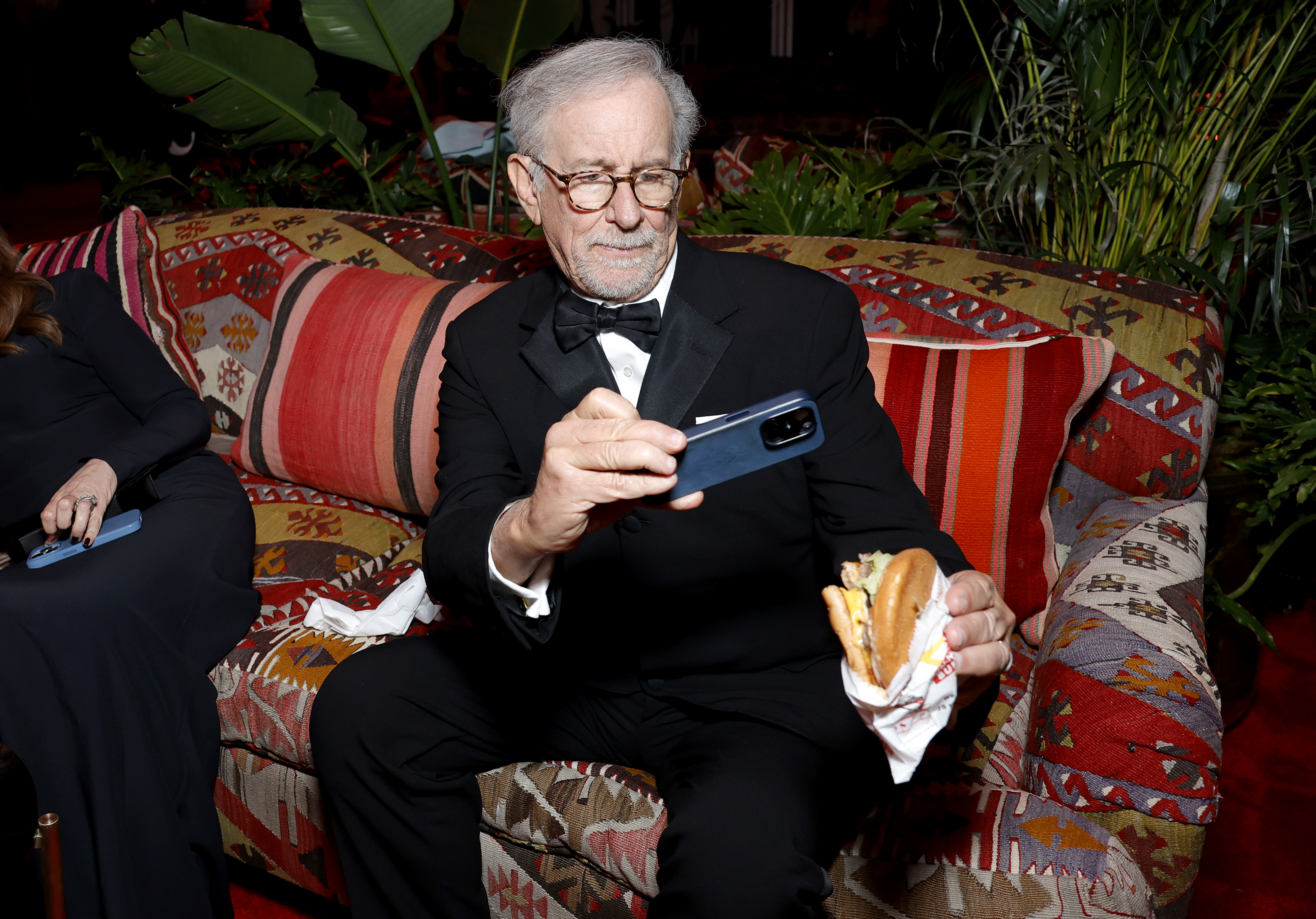 Steven Spielberg in a tuxedo, sitting and taking a pic of his burger