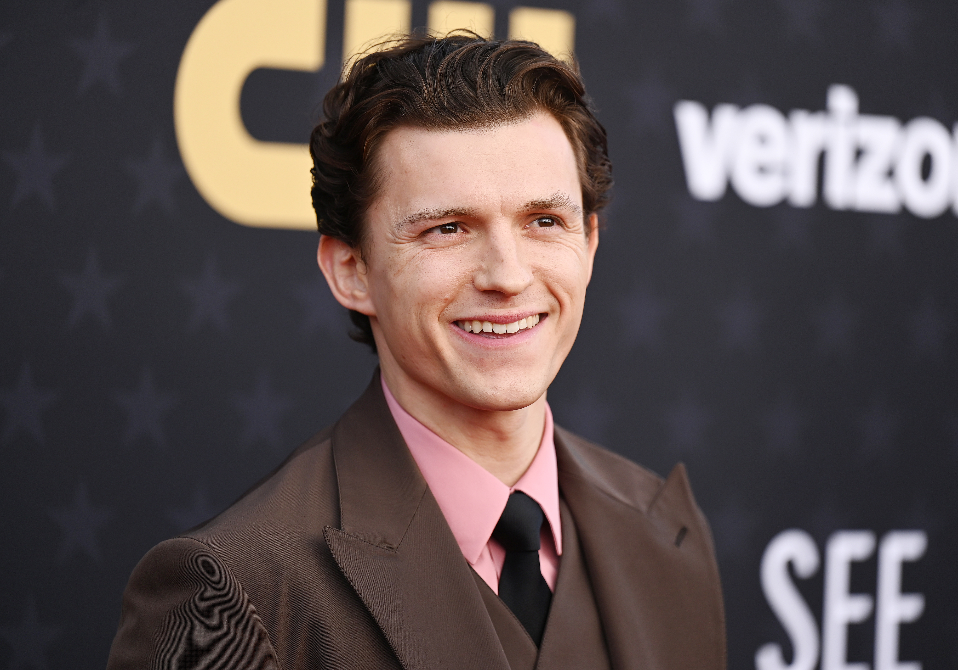 Tom Holland in a brown suit with a pink tie, smiling on the red carpet