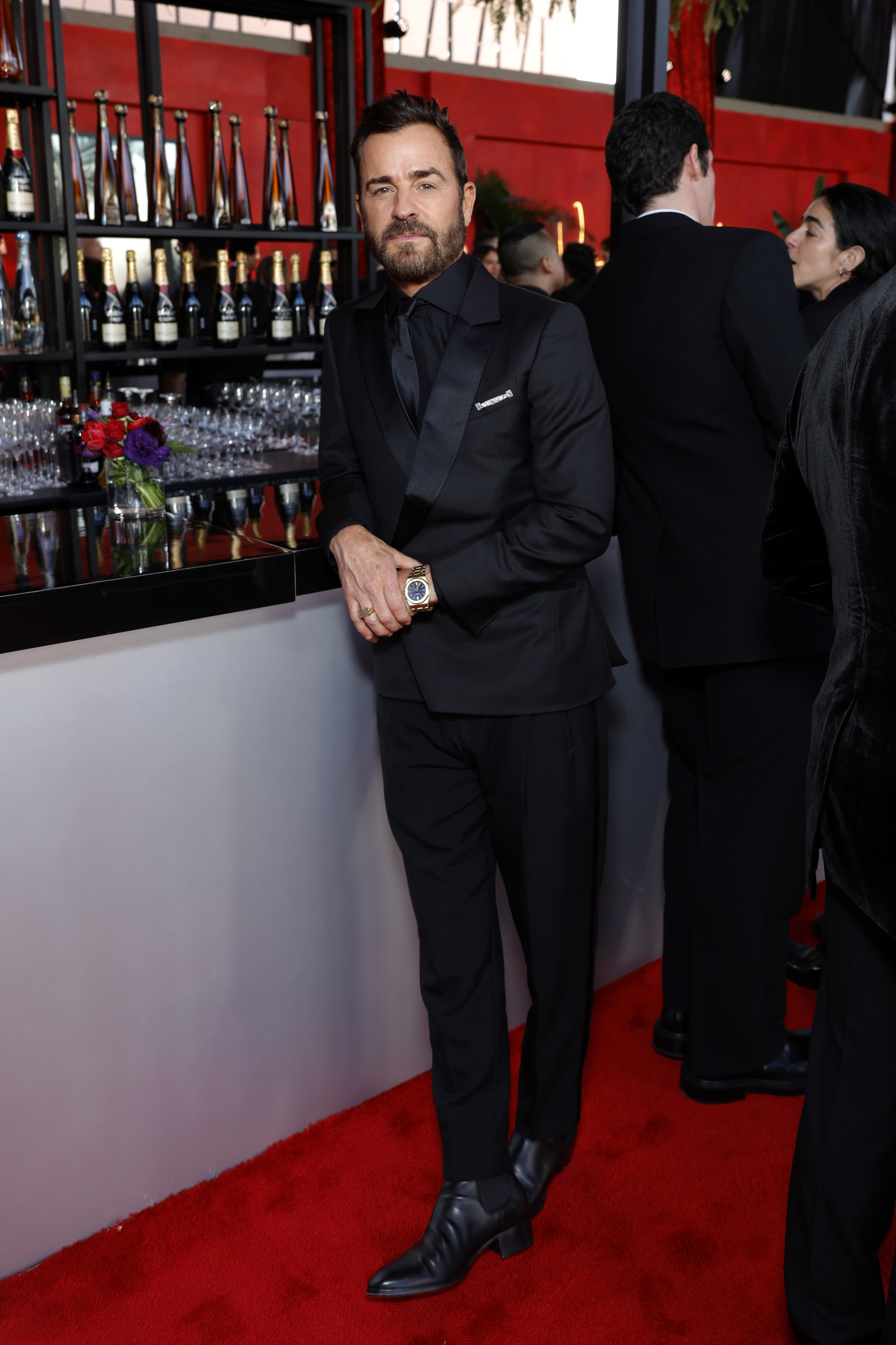 Justin Theroux in a black suit, standing on a red carpet, leaning against a bar with bottles in the background