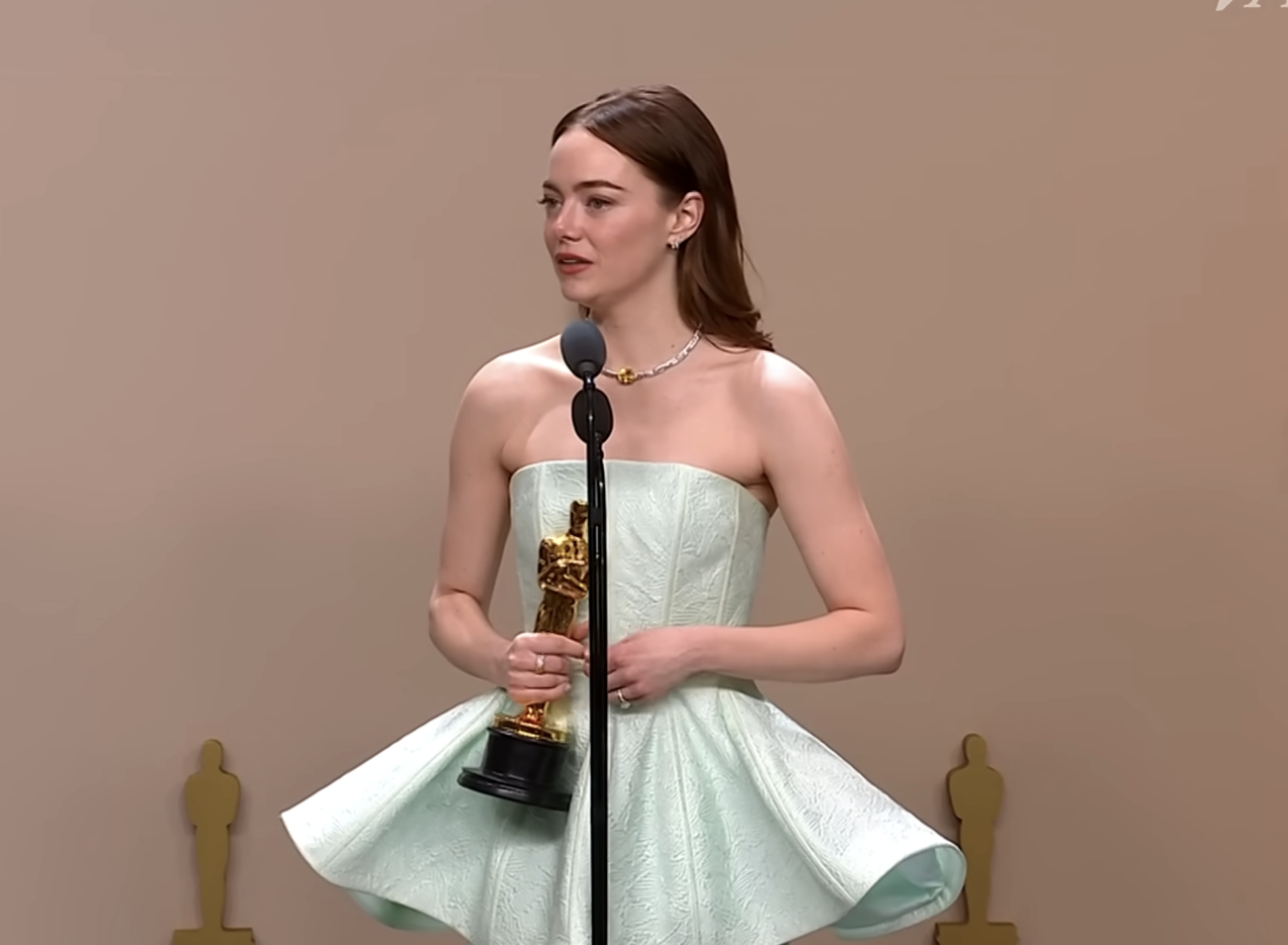 Emma Stone in a full-sleeved, high-neck gown stands with a microphone in front of Oscar statues