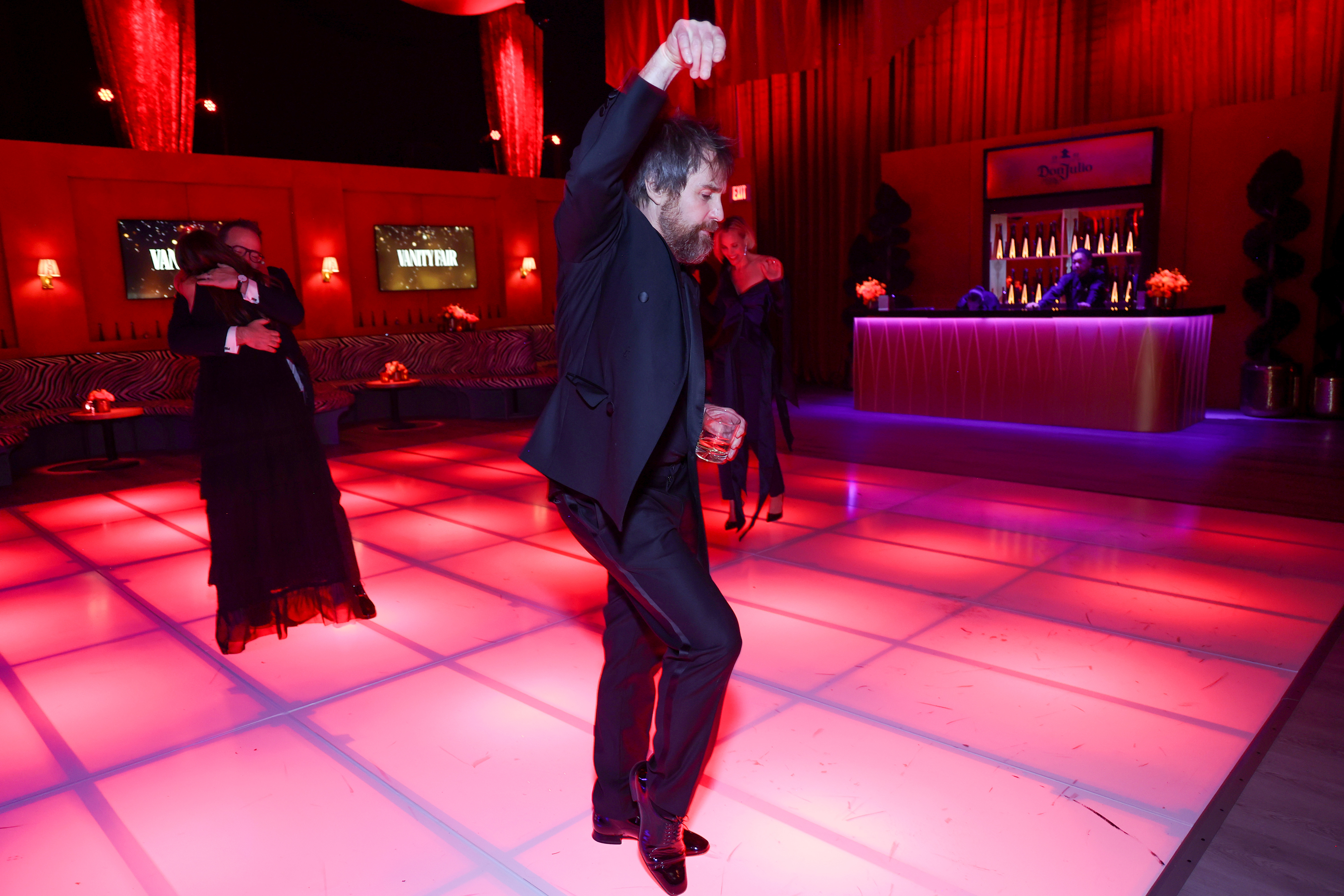 Sam in a suit dancing with a raised fist on a lit dance floor, other couples in background