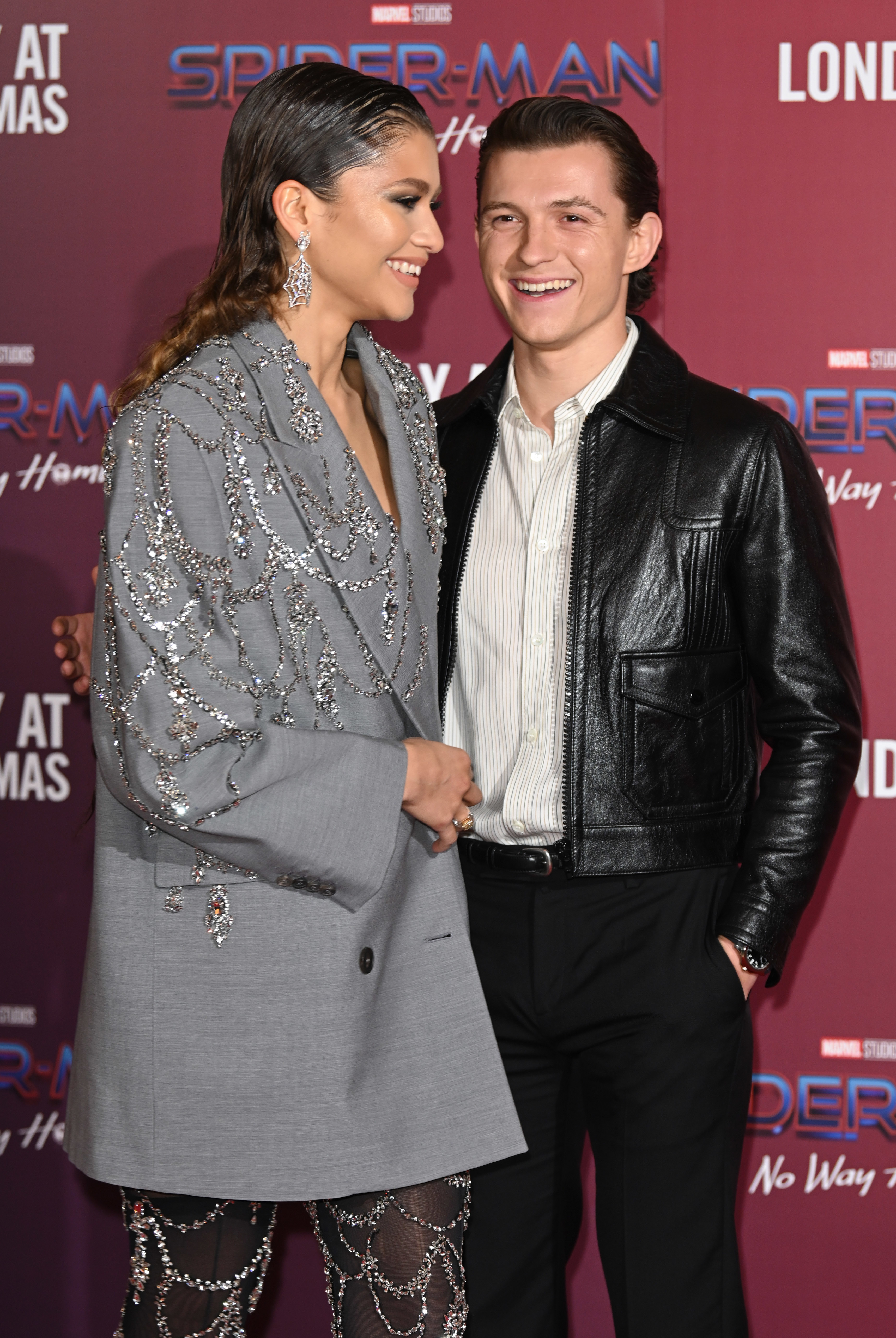 Zendaya in a long beaded suit dress and Tom in a leather jacket and striped shirt at a media event