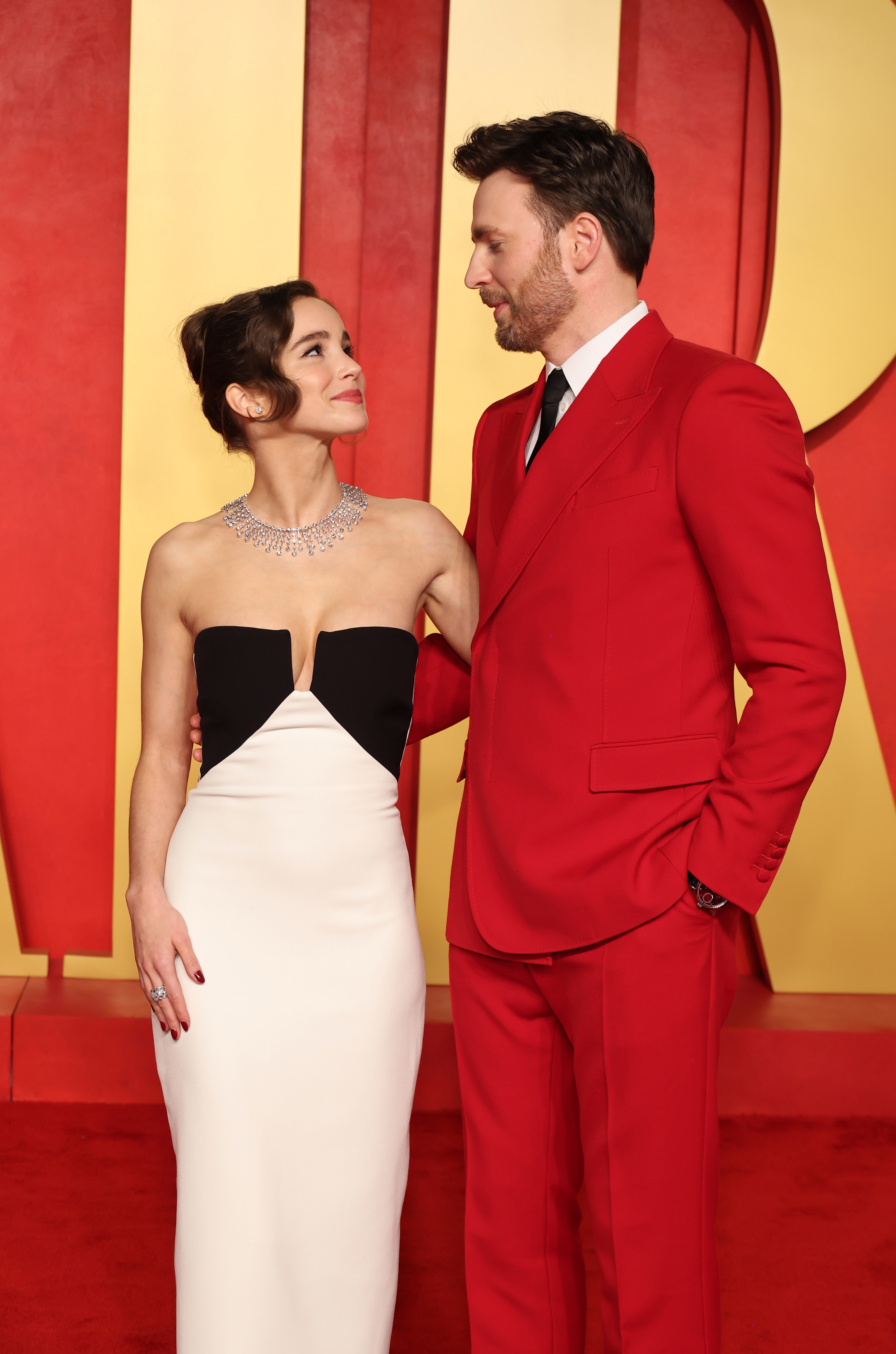 Alba in a white dress with a black top, Chris in a red suit, looking at each other