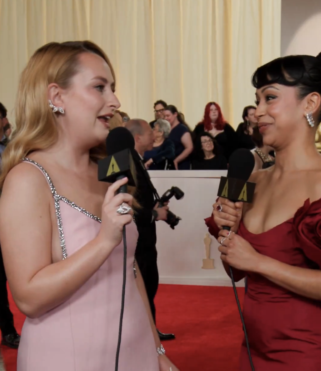 The two wo women interviewing on red carpet, Amelia in crystal-embellished gown, Liza in off-shoulder ruched dress