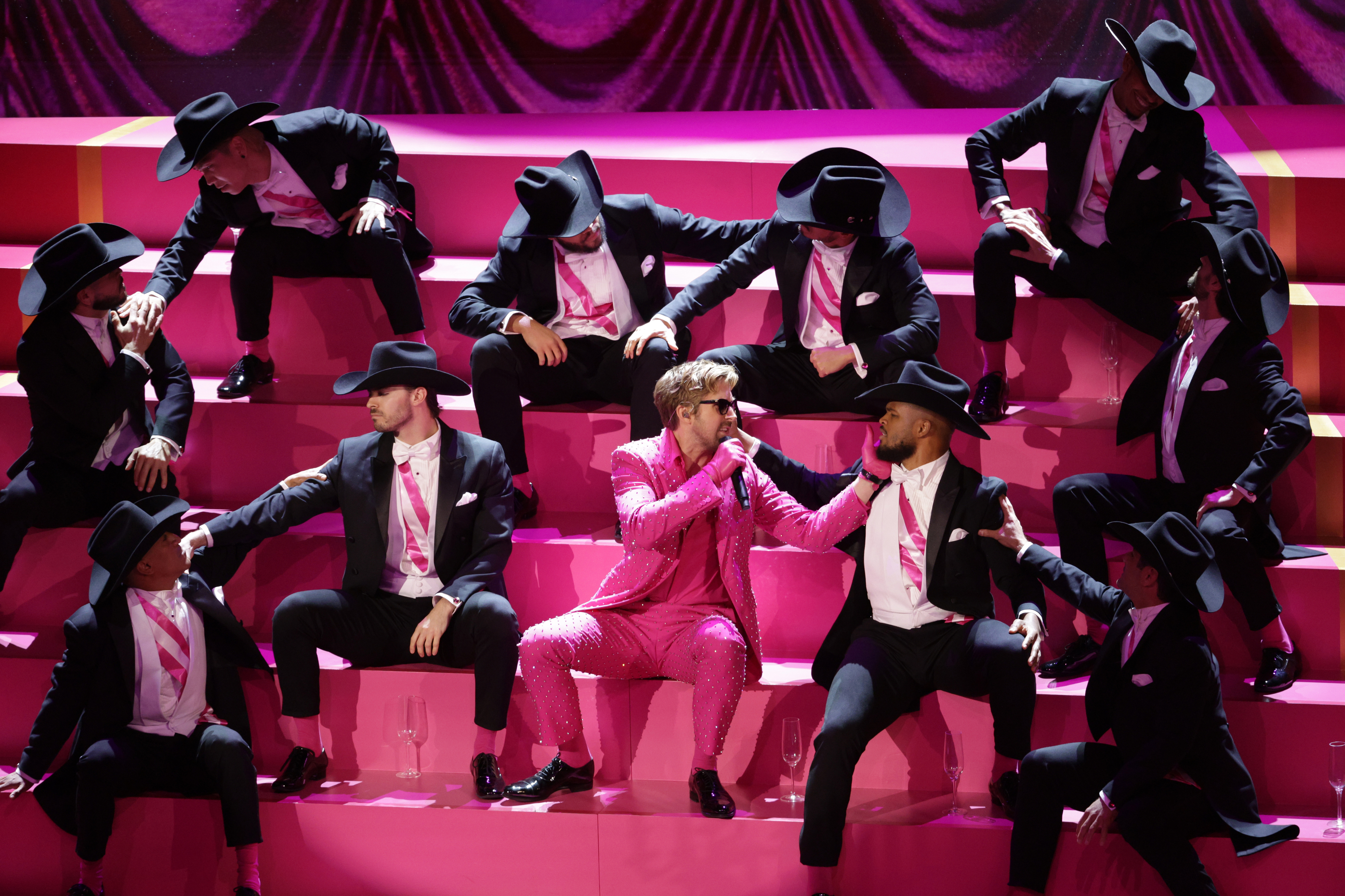 Ryan in a pink suit at the center holding a microphone, with other performers in pink and black attire sitting on steps onstage