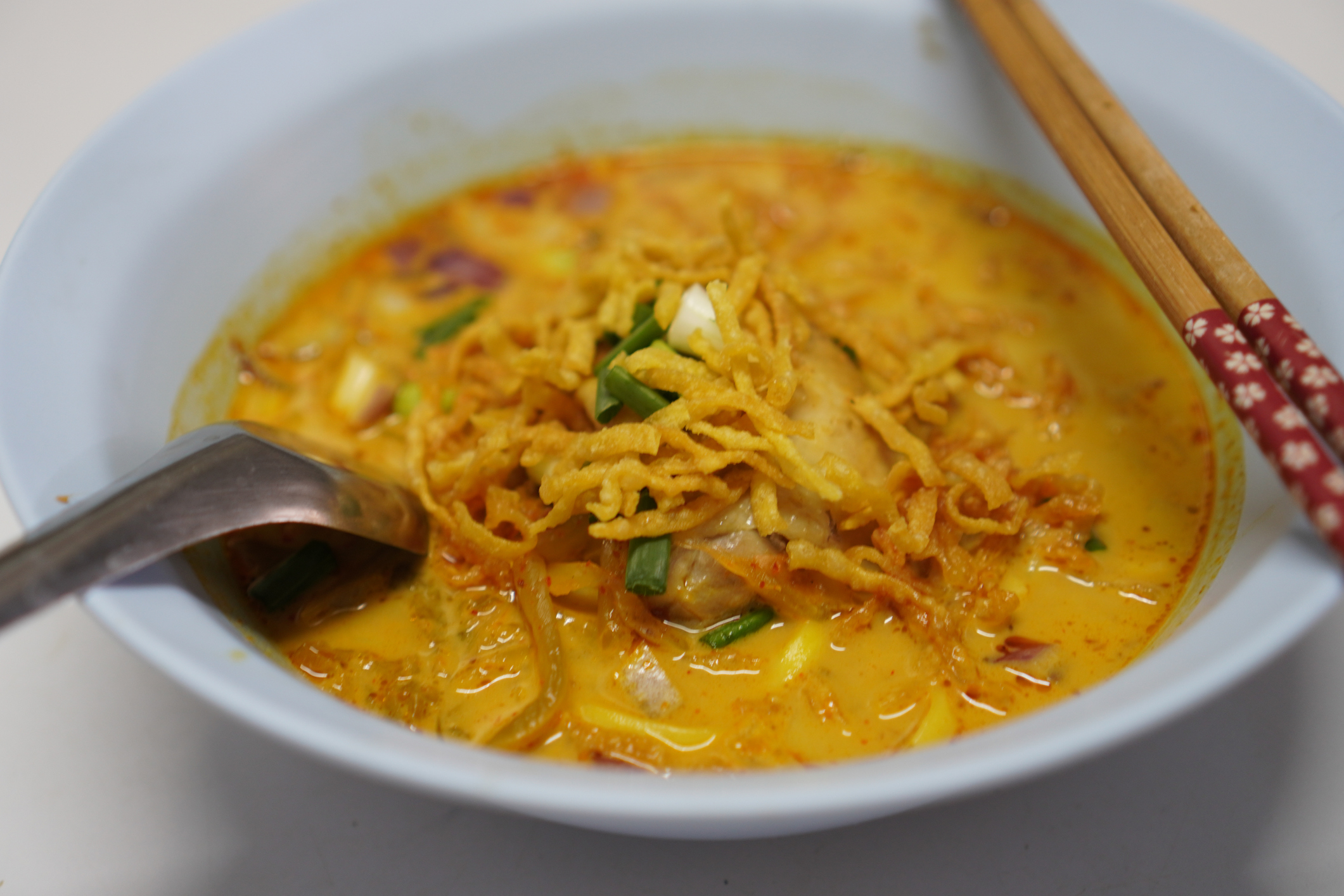 A bowl of khao soi with spoon and chopsticks, featuring noodles and green onions