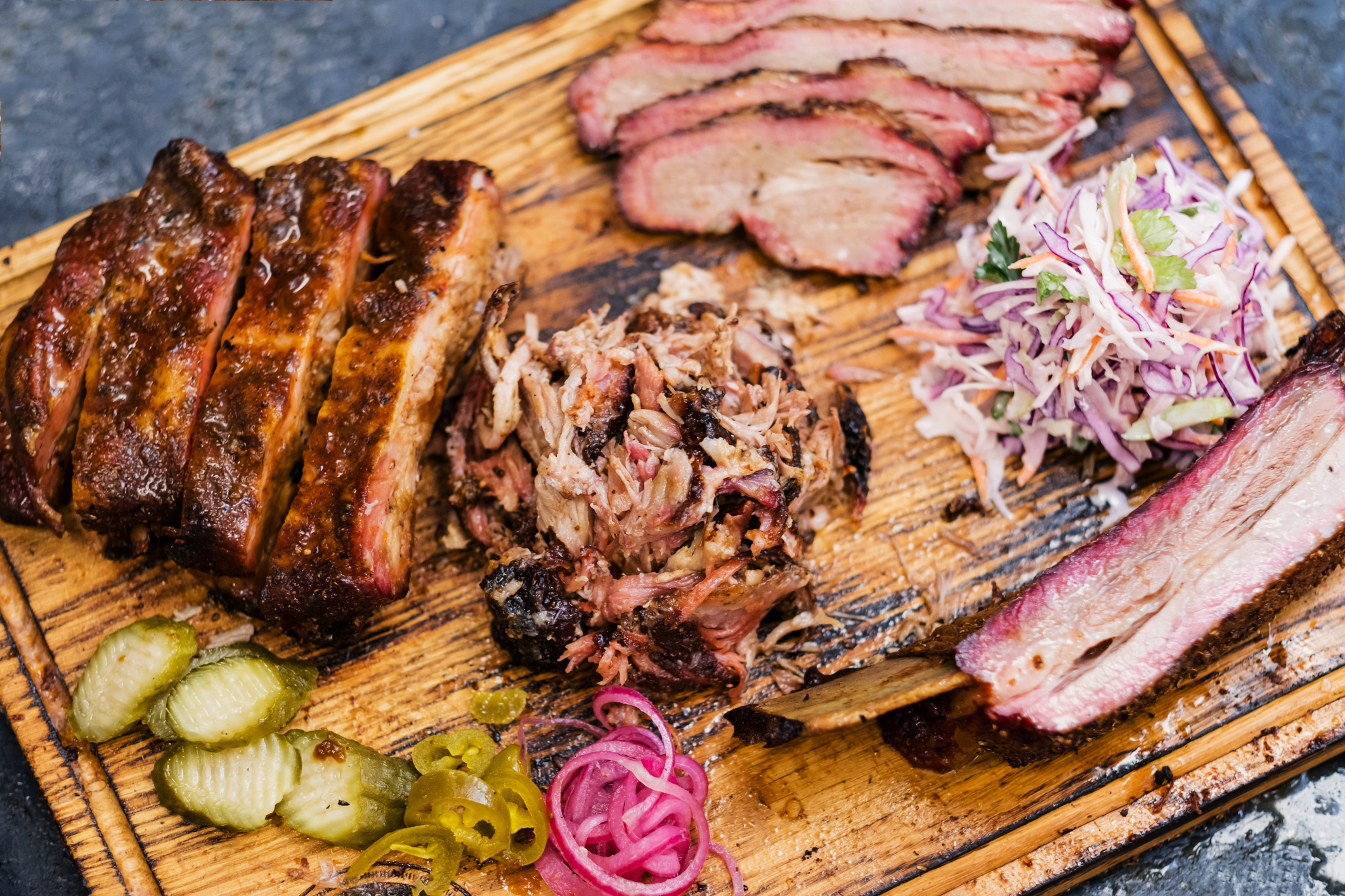 Smoked meat on a wooden board with ribs, pulled pork, brisket, coleslaw, pickles, and onions
