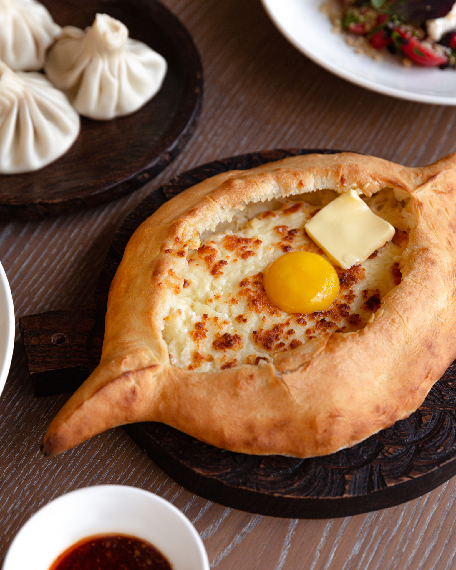 Georgian Khachapuri: bread boat with melted cheese, butter pat, raw egg on top, side of dumplings